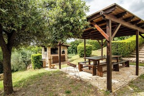 Ein Außenbereich mit einer Terrasse, einem Steintisch und -bänken, einer Holzpergola und einem Kamin aus Stein.