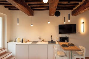 A modern kitchen with a wooden island, white cabinetry, pendant lights, a flat-screen TV, and a dining area with white chairs.