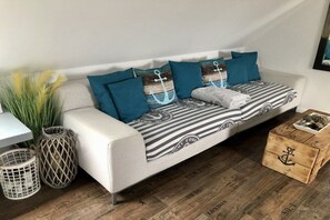 A white sofa with striped and anchor-themed cushions, a wooden crate with an anchor design, and two wicker baskets on a wooden floor. - Anchorage (To the old coach house Norddeich) (Norddeich)