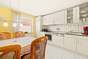 Een compacte keuken met witte kastjes, een betegelde spatwand en een eethoek met een gestreepte tafelkleed en patroonstoelen.