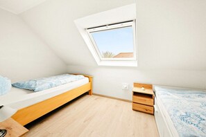 Een kleine, goed verlichte kamer met een schuine kap, dakraam, twee eenpersoonsbedden, een houten bureau en een klein dressoir.