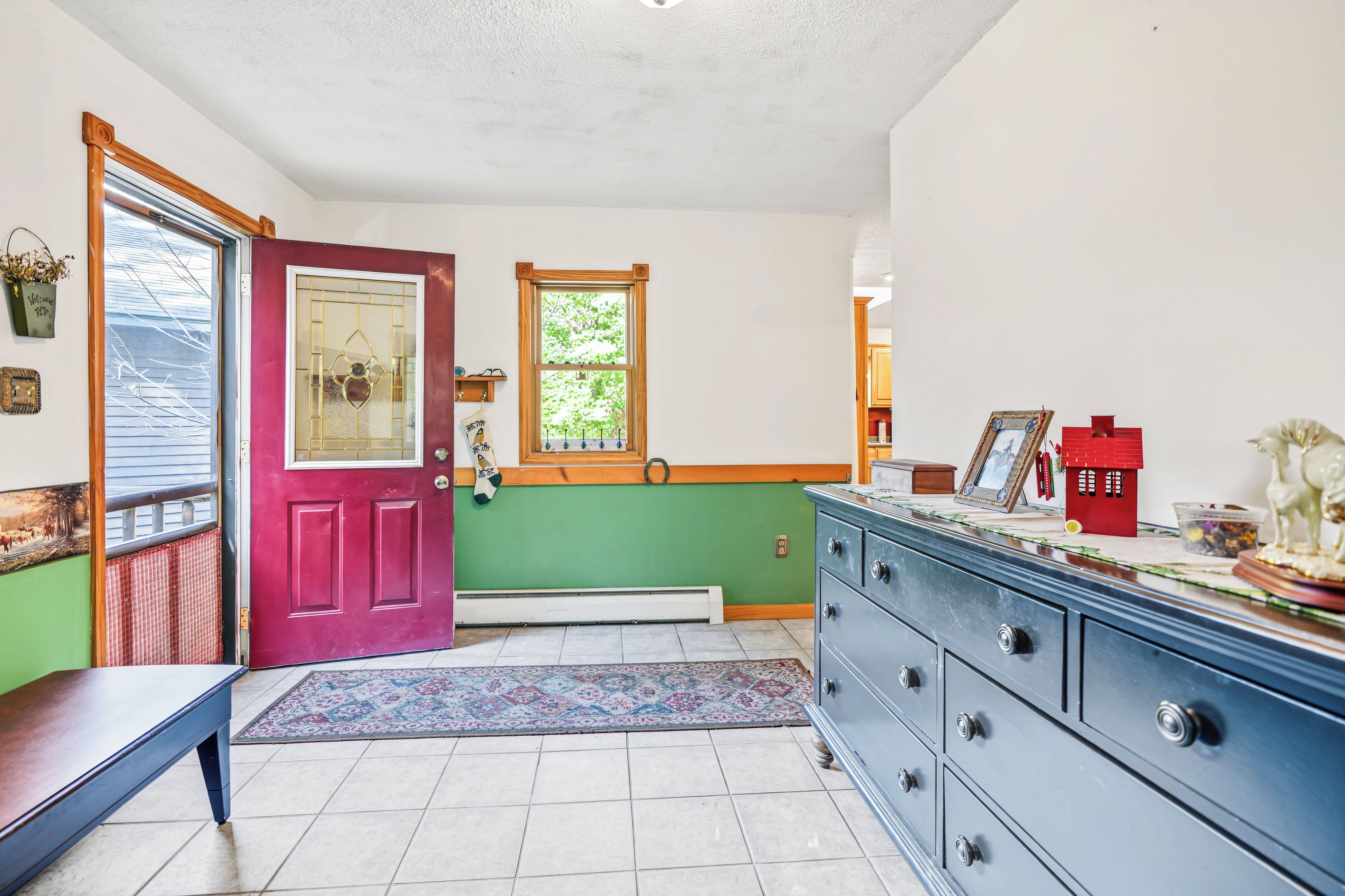 A room with a red door, a window with a view of trees, a blue dresser, and a patterned rug.