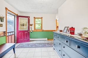 A room with a red door, a window with a view of trees, a blue dresser, and a patterned rug. - Little Camp Christmas (Plattsburgh)