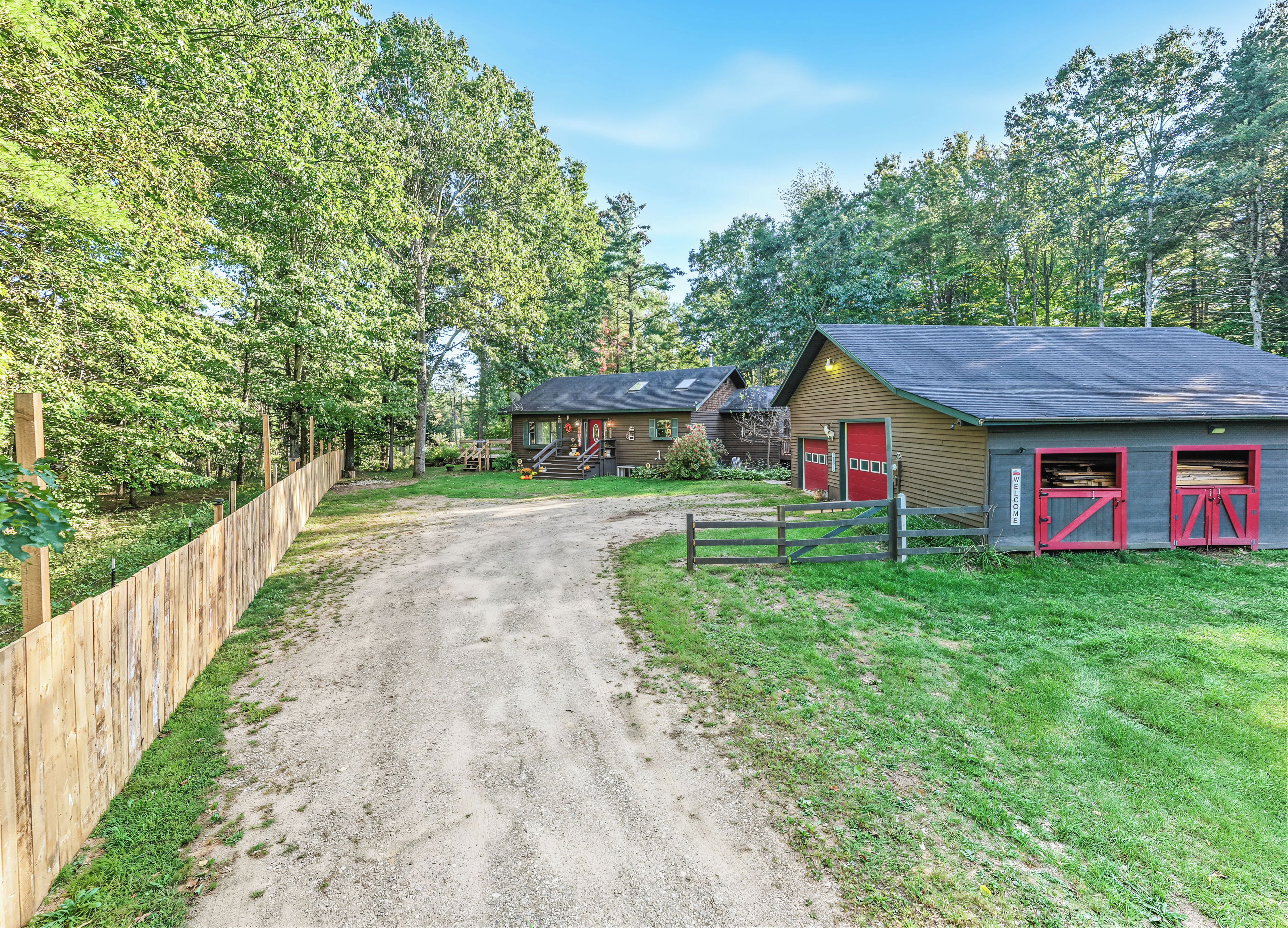 A rural property with a gravel driveway leading to a wooden fence, a barn with red doors, and a small house with a dark roof.