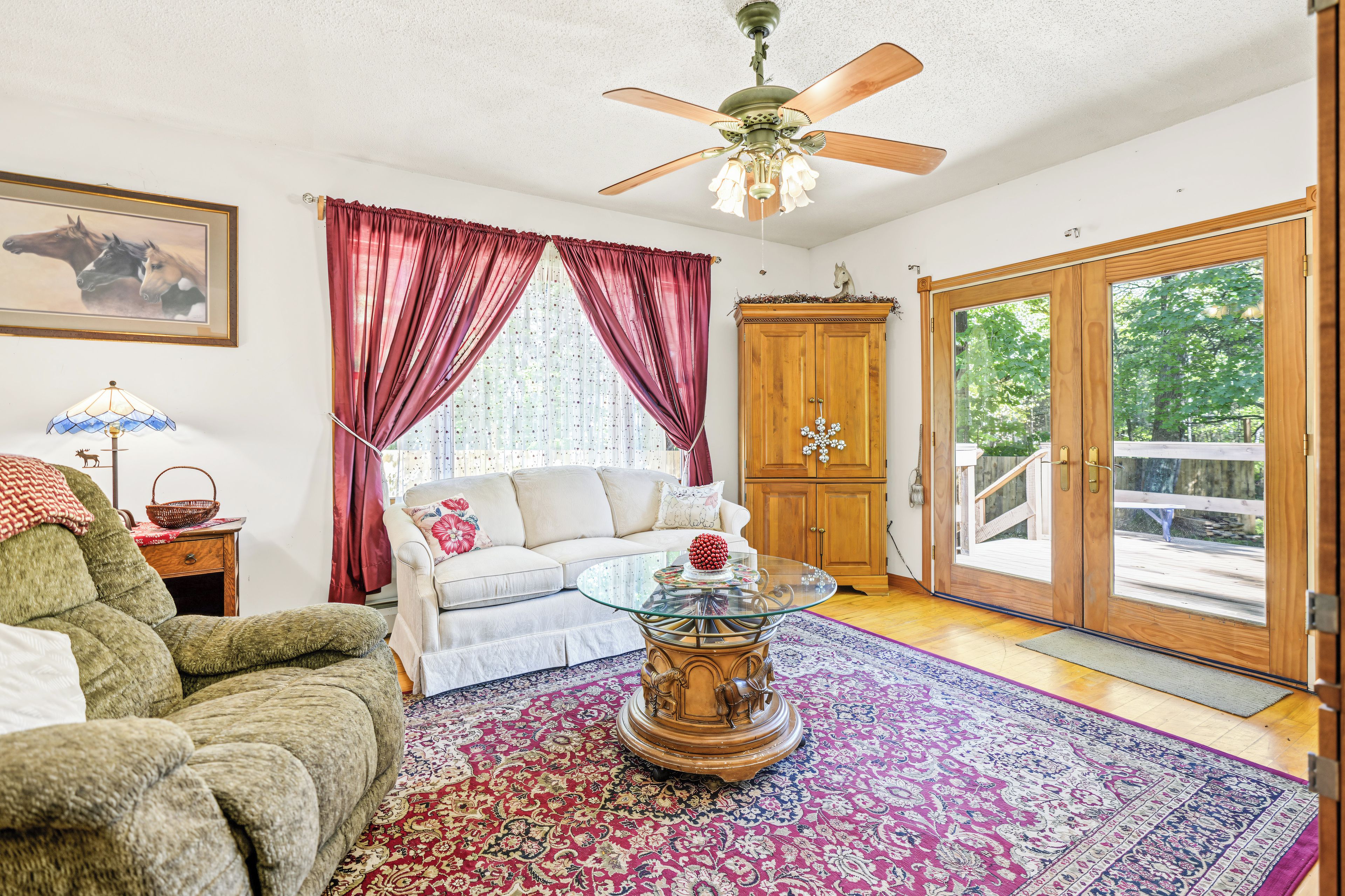 A living room with a sofa, a glass coffee table, a red and white patterned rug, a wooden cabinet, and a ceiling fan.
