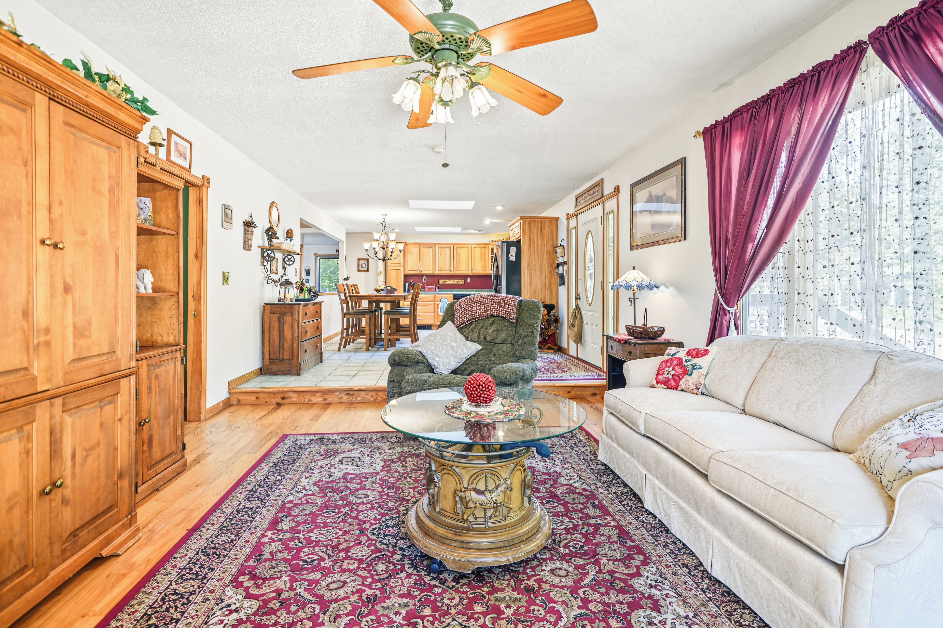A spacious living room with a ceiling fan, a dining area, wooden cabinets, and a large window with curtains.