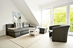 A modern living room with a sofa, armchair, and coffee table, large windows with a view of greenery, and a sloped ceiling. - Frisian happiness on the dike (Norddeich)