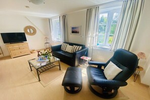 Uma sala de estar com sofá, poltrona, mesa de centro, televisão e janela com cortinas.