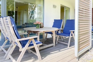 Eine Holzterrasse mit einem runden Tisch und vier blauen Stühlen, eine Glastür zum Innenbereich und ein Blumentopf mit rosa Blüten.