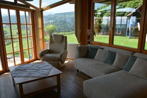 Ein Zimmer mit einer Couch, einem Sessel und einem Tisch. Große Fenster bieten Blick auf einen Garten und Hügel.