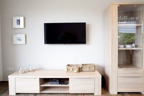 A modern living room with a flat-screen TV mounted on the wall, a wooden TV stand with baskets, and a glass cabinet with dishes. - Holiday apartment with 2 bedrooms (Norddeich)