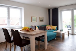 A modern living room with a wooden dining table, a teal sofa, and a large window providing natural light. - Holiday apartment with 2 bedrooms (Norddeich)