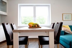 A dining area with a table and chairs, a fruit bowl on the table, a window with a view of greenery, and a teal sofa in the background. - Holiday apartment with 2 bedrooms (Norddeich)