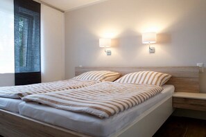 A double bed with striped bedding, two wall-mounted light fixtures, a wooden headboard, and a nightstand with a drawer. - Holiday apartment with 2 bedrooms (Norddeich)