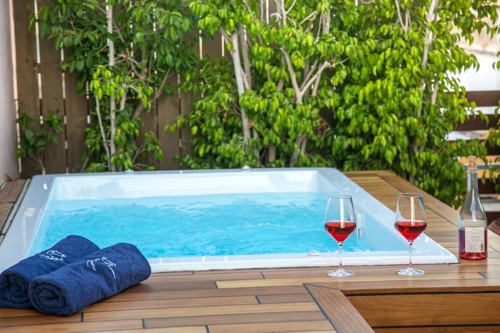A hot tub with two glasses of red wine and a bottle of wine on a wooden deck.