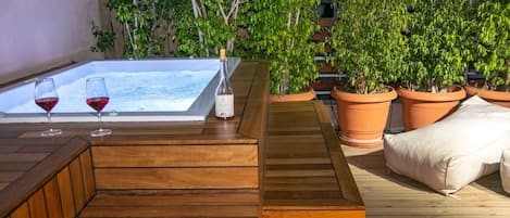 Una terraza de madera con un jacuzzi, dos copas de vino tinto y macetas con plantas.