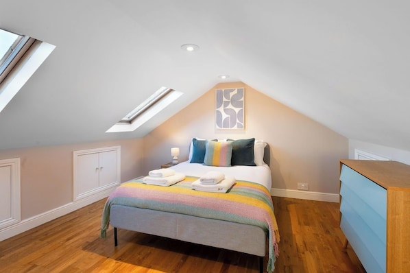 Un dormitorio con techo inclinado, una cama con manta de colores de arcoíris, una mesita de noche con lámpara y un aparador de madera.