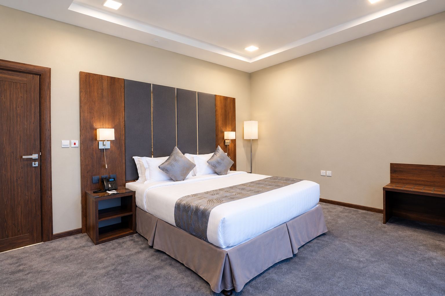 A hotel room with a large bed, bedside tables, a desk, and a bench.