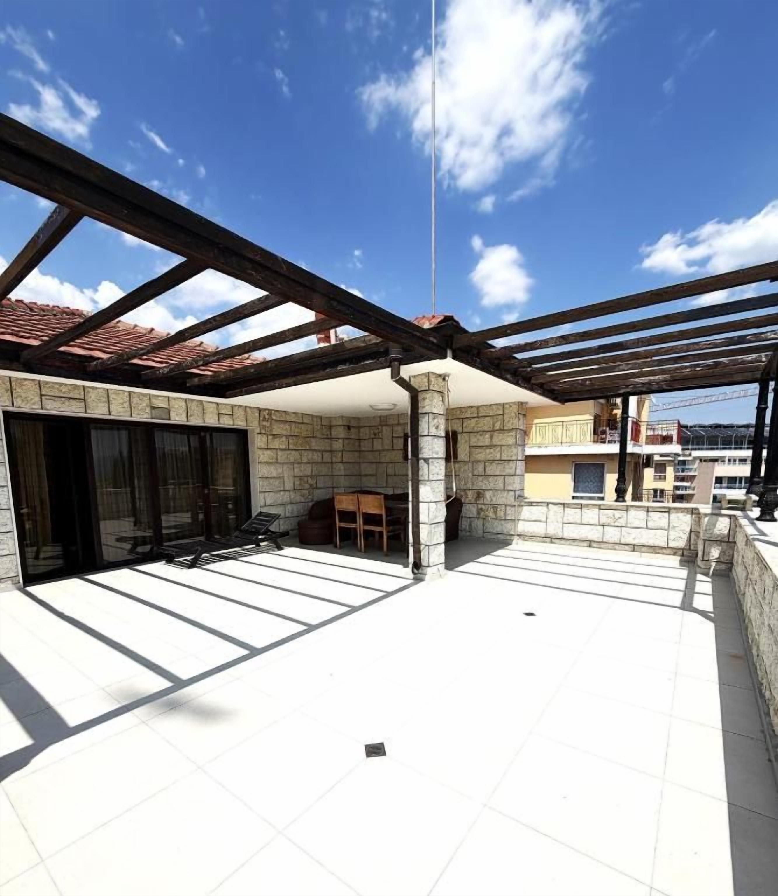 Área externa coberta com parede de pedra, pérgola de madeira e espaço de estar com cadeiras e mesa.