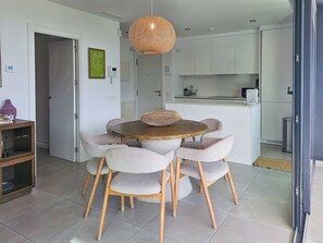 Ein Essbereich mit einem runden Holztisch, vier gepolsterten Stühlen, einer geflochtenen Pendelleuchte und einer Küche im Hintergrund.