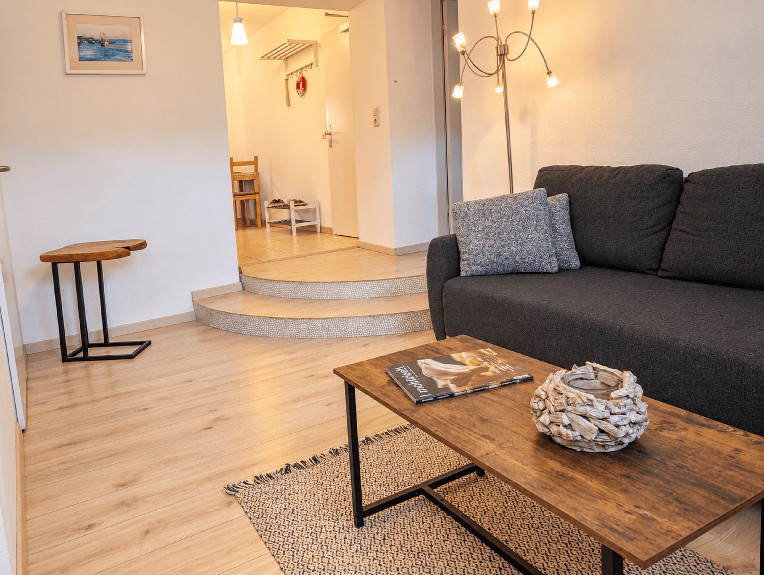 Una sala de estar moderna con un sofá, una mesa de centro con un libro y un cuenco decorativo, un taburete de madera pequeño y una escalera que conduce a otra habitación.