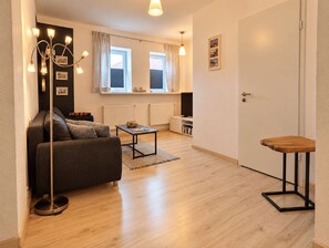 A modern living room with a sofa, coffee table, and a small wooden stool. There is a radiator, a television, and a window with curtains. - Nordnest: Holiday apartment in the center of Kappeln with parking and bike shed (Kappeln)