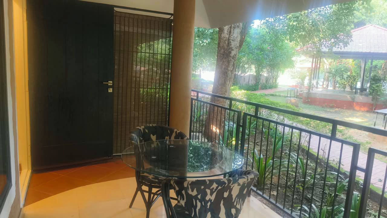Un balcón con mesa y sillas de vidrio, vista a un jardín y una puerta con mosquitero de metal.