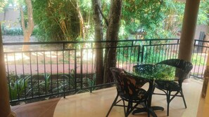 Un balcone con tavolo e sedie, circondato da una ringhiera metallica, affacciato su un giardino con alberi e piante.