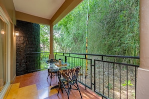 A balcony with a glass table and chairs, a stone wall, and a view of dense greenery.