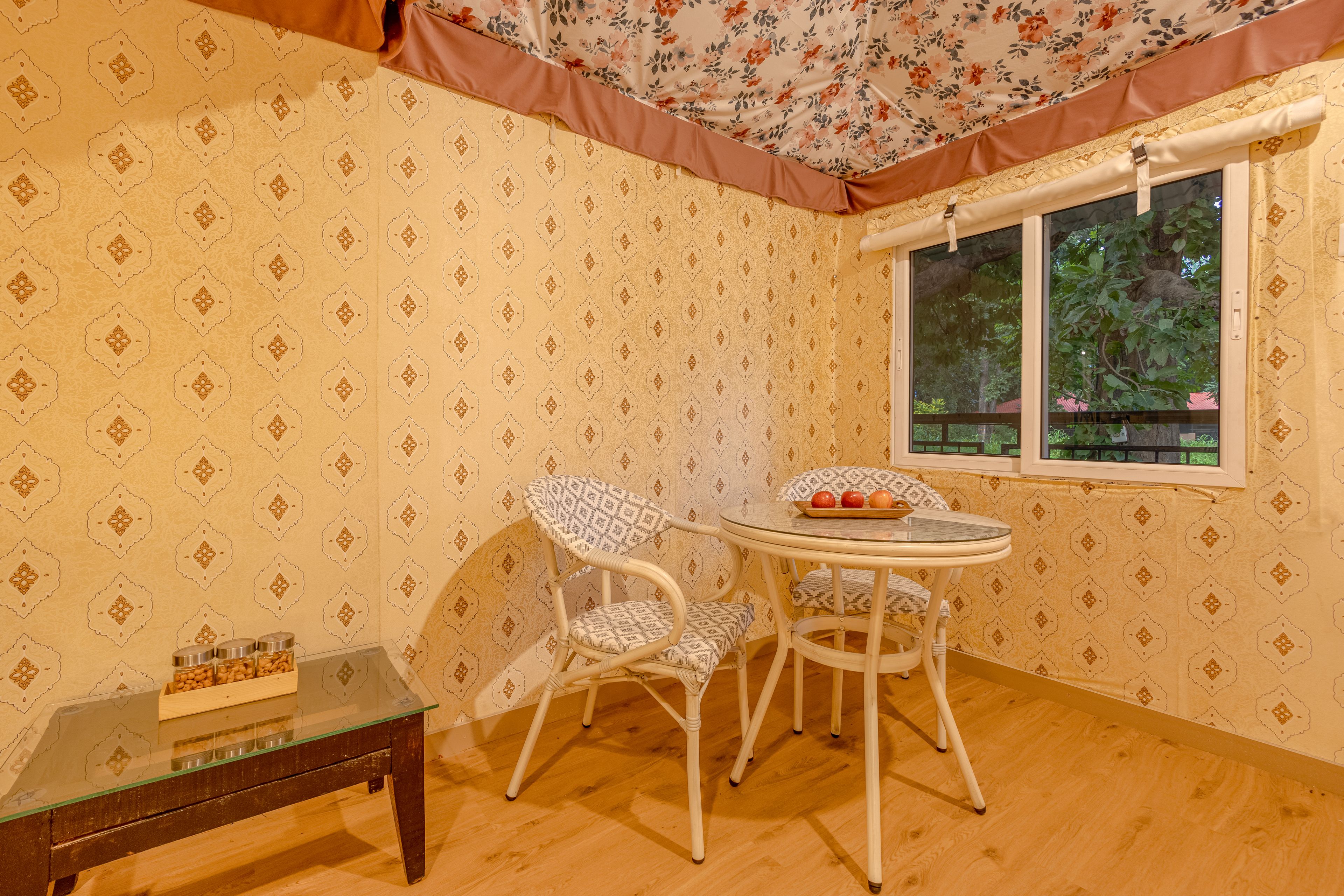 Ein Zimmer mit Wandtapete mit Muster, einem runden Glastisch, zwei weißen Stühlen, einem kleinen Holztisch mit Gläsern und einem Fenster mit Blick auf Bäume.