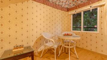 Ein Zimmer mit Wandtapete mit Muster, einem runden Glastisch, zwei weißen Stühlen, einem kleinen Holztisch mit Gläsern und einem Fenster mit Blick auf Bäume.