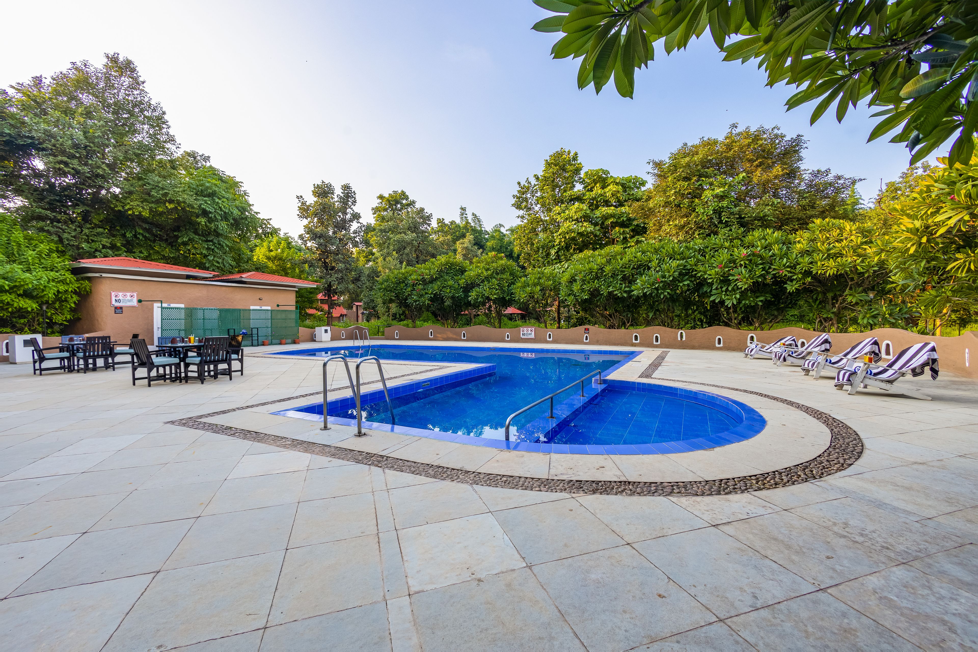 Una piscina amb zona de solàrium, envoltada de vegetació i un edifici amb teulada vermella.