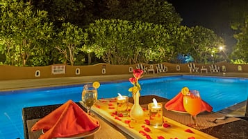 A poolside table with a bottle, glasses, and a candle, surrounded by greenery and a pool.