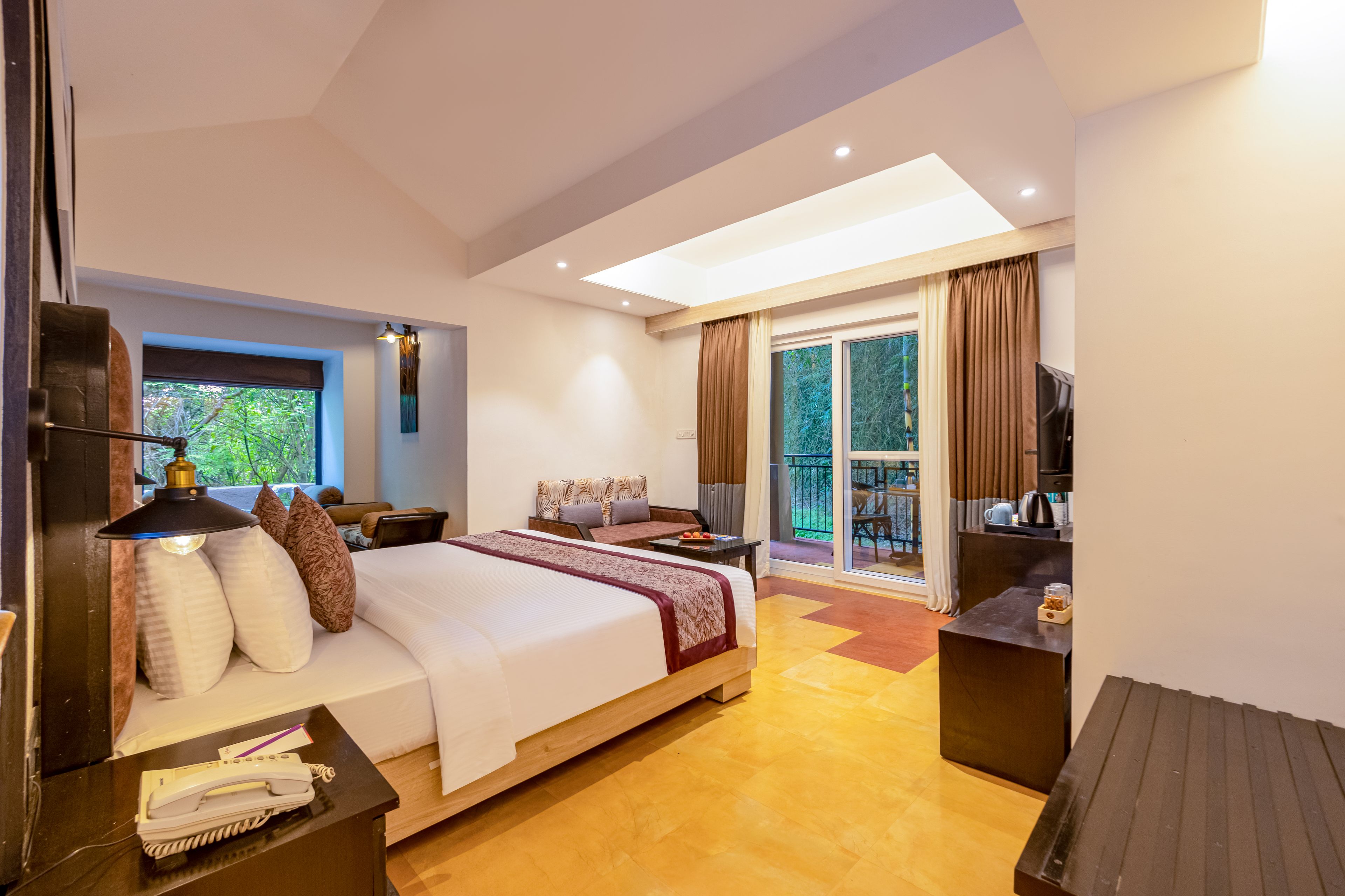 A modern hotel room with a large bed, a desk, a chair, a TV, and a balcony with a view of trees.
