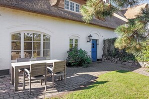 Een wit huis met een blauwe deur, een rieten dak en een terras met tegels, een tafel en stoelen.