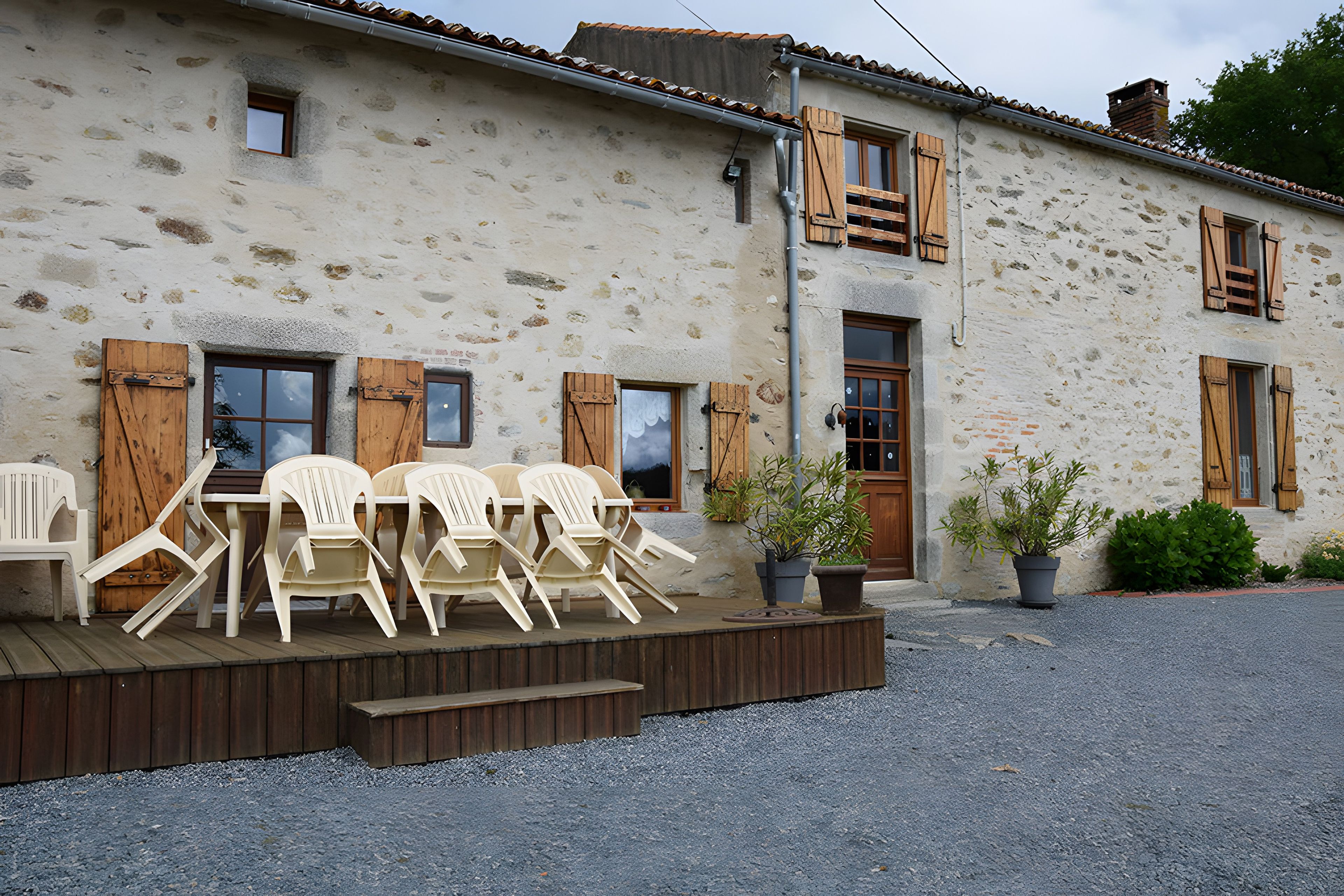 A rustic stone building with wooden shutters, a terrace with plastic chairs, and potted plants.