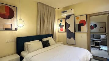 A hotel room with a bed, a bedside table, a chair, a wall-mounted air conditioner, and abstract art on the walls.
