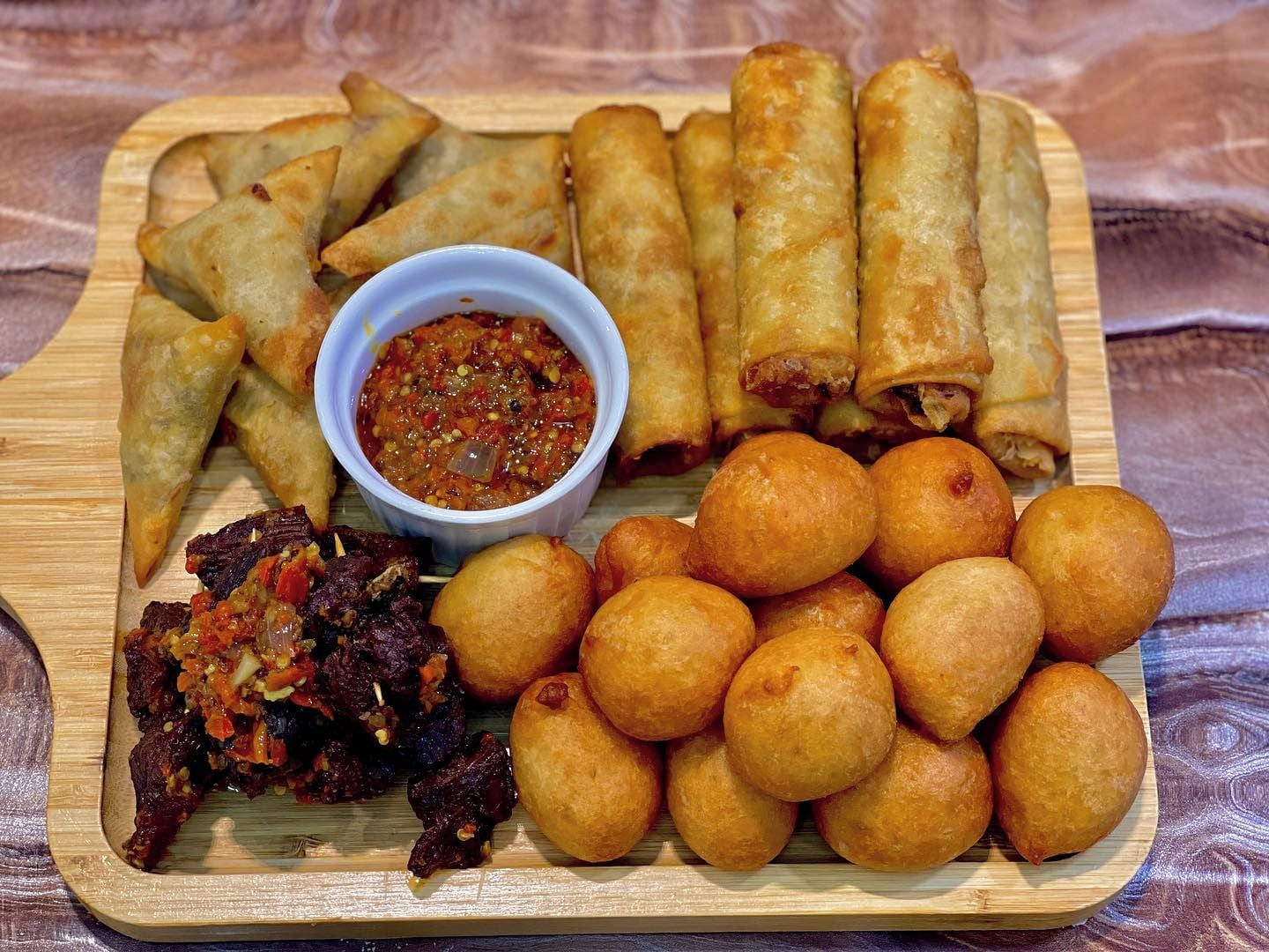 A wooden tray with various fried snacks, including samosas, spring rolls, and fried dough balls, accompanied by a bowl of chutney.