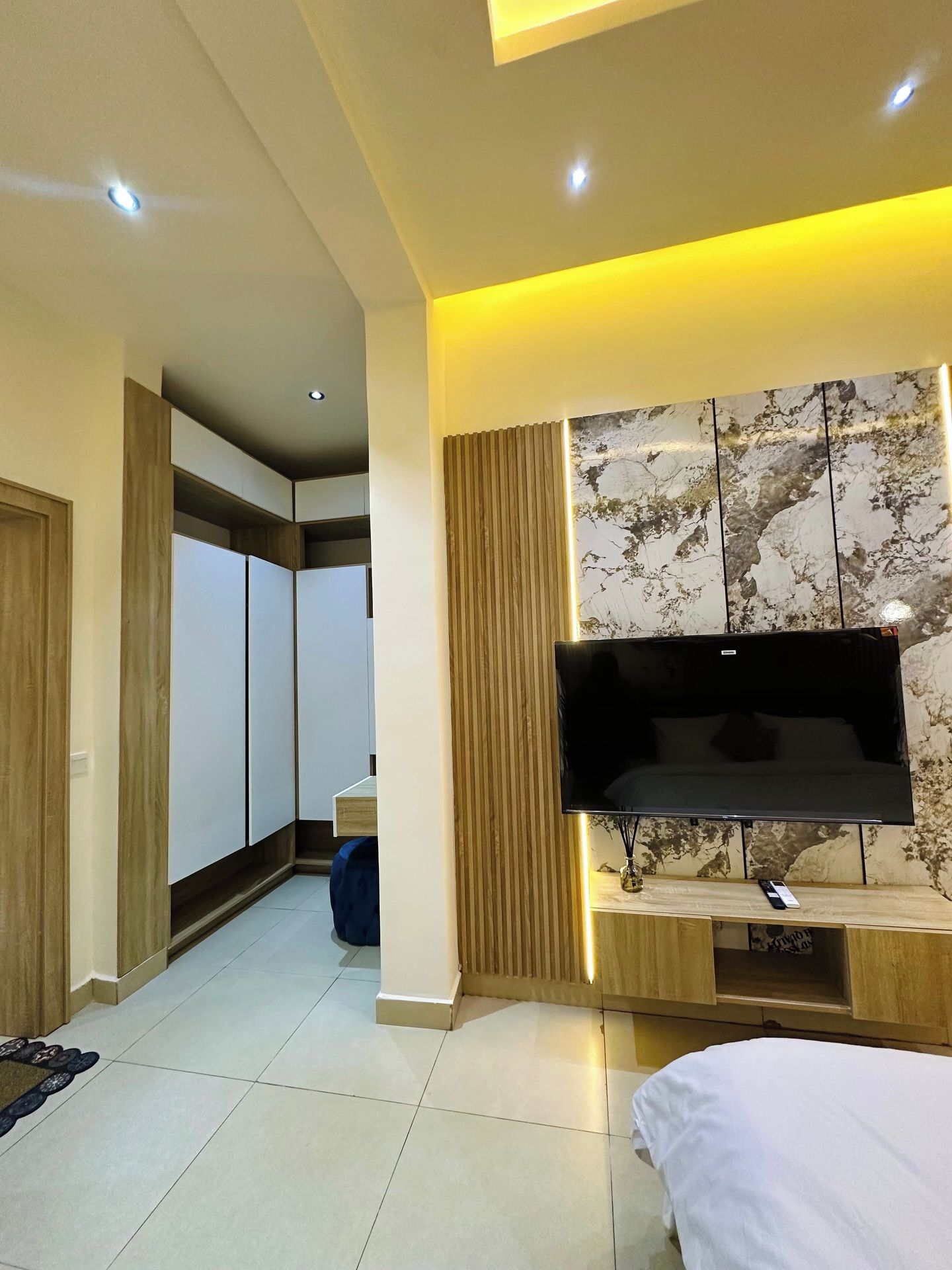Una habitación de hotel moderna con una televisión de pantalla plana montada en un panel de madera, un espejo grande y una cama con cubrecama blanco.