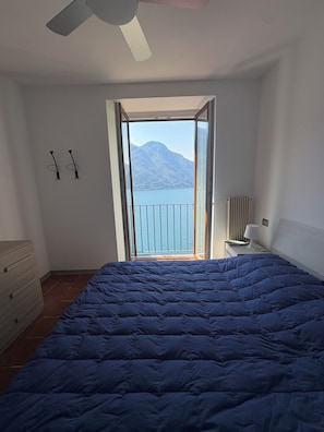 Un dormitorio con cama, cómoda, mesita de noche, lámpara y vistas a una montaña y lago a través de una puerta abierta.