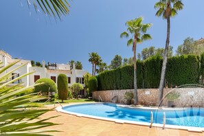 Une piscine bordée d’un mur en pierre et entourée de palmiers, le tout agrémenté d’un jardin bien entretenu et d’un bâtiment doté d’un toit en tuiles.