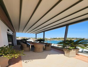 Eine Terrasse im Freien mit Esstisch und Stühlen, große Fenster mit Meerblick und Blumentöpfen.