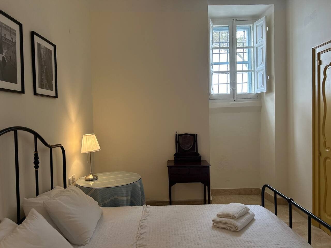 A bedroom with a bed, a bedside table with a lamp, a chair, a window, and a framed picture on the wall.