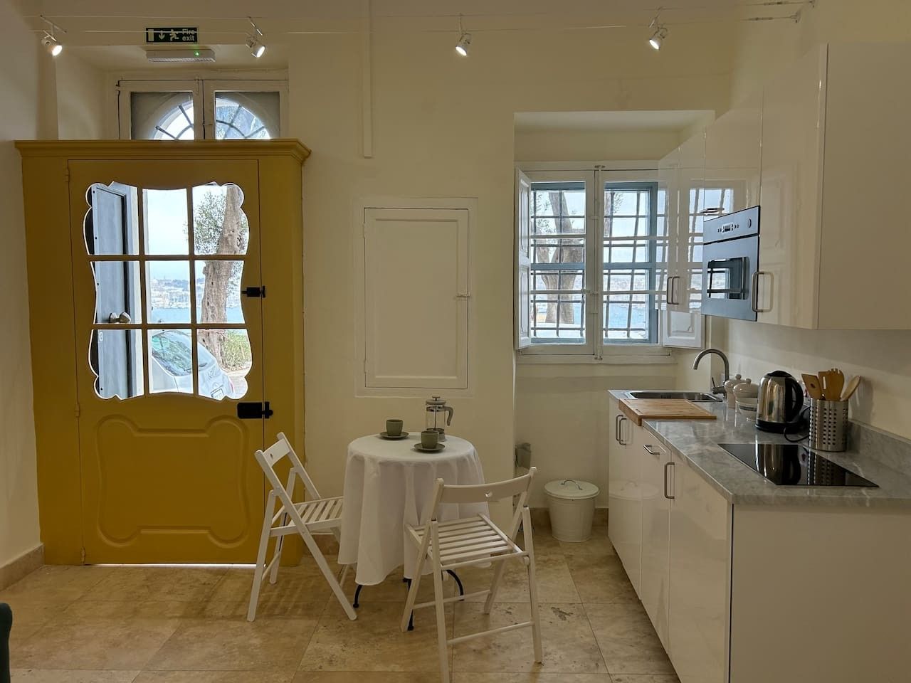A compact kitchen with a dining area, a yellow door with a window, a white table with chairs, and a sink.