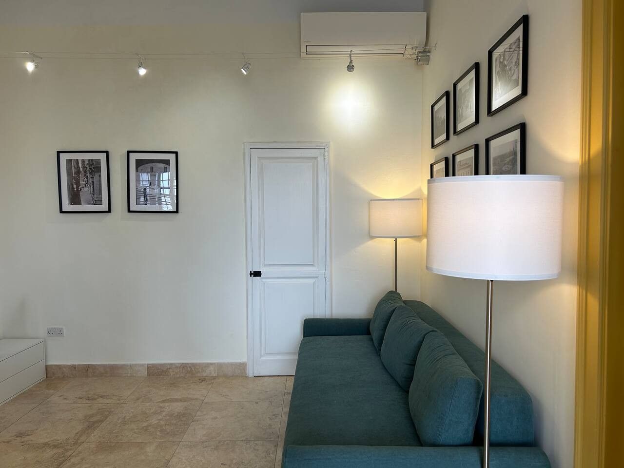Een kamer met een smaragdgroene bank, een staande lamp en ingekaderde foto's aan de muur.