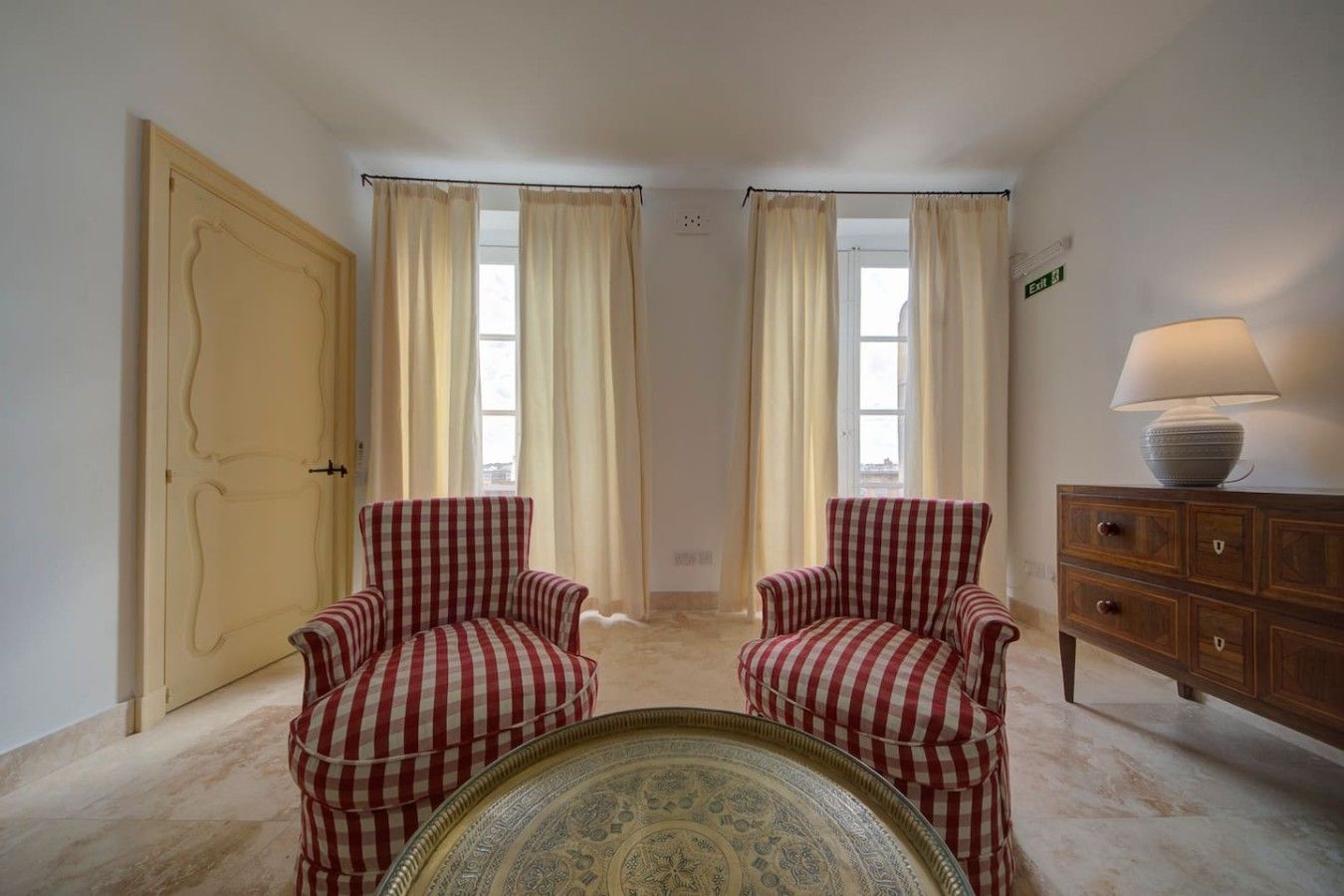 A room with two checkered armchairs, a wooden cabinet with a lamp, and a patterned table.