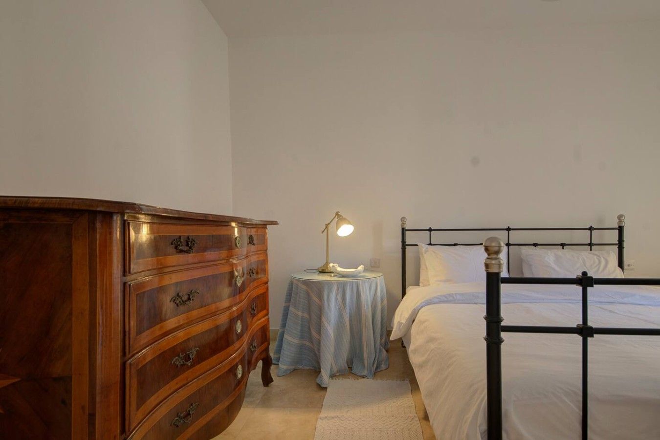 A bedroom with a wooden dresser, a round bedside table with a lamp, and a metal-framed bed.