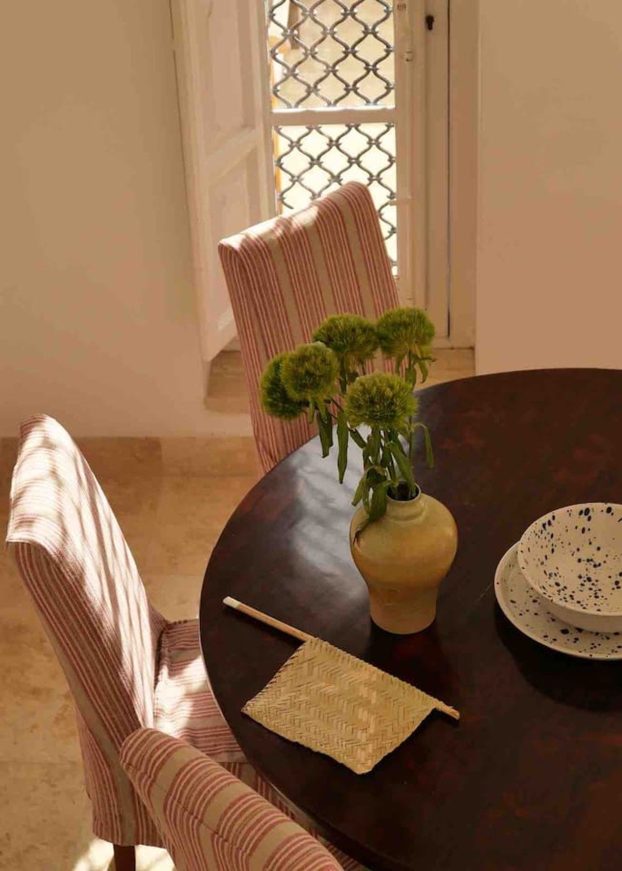 A dining table with a vase of green plants, a woven placemat, and a bowl with speckles.