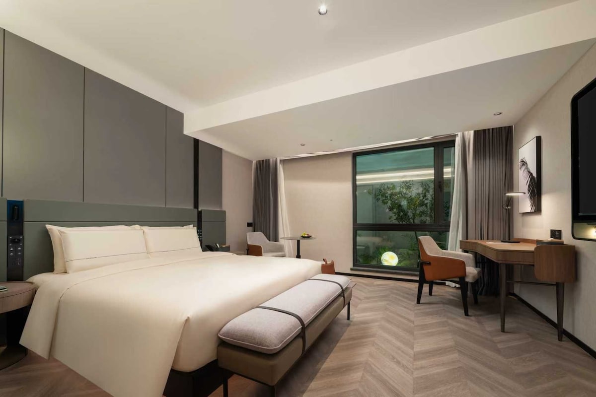 Habitación de hotel moderna con una cama grande, un escritorio, una silla y vistas a zonas verdes a través de una ventana.