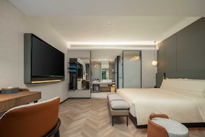 Habitación de hotel moderna con televisión de pantalla plana, escritorio, cama, banco y baño con espejo y lavamanos.